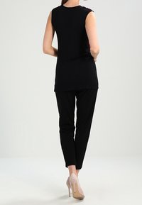 Black sleeveless top with a curved neckline and solid back, paired with black tapered trousers and light-colored high-heeled shoes.