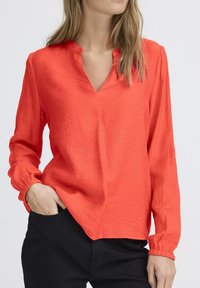 Halenka s výstrihom do V a dlhými rukávmi v jasne oranžovej farbe, vyrobená z textúrovanej látky. Disponuje elastickými manžetami a voľným strihom, skombinovaná s tmavými nohavicami.