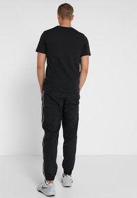 Camiseta negra de manga corta combinada con pantalones negros que tienen franjas blancas a los lados. El modelo está de pie con las manos en los bolsillos, mostrando el ajuste.