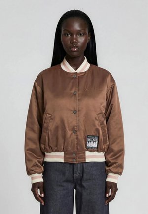 Frau mit langen, glatten Haaren, trägt eine braune Satin-Bomberjacke mit weißen, gerippten Bündchen und Kragen sowie dunkelblaue Jeans, vor einem schlichten Hintergrund.