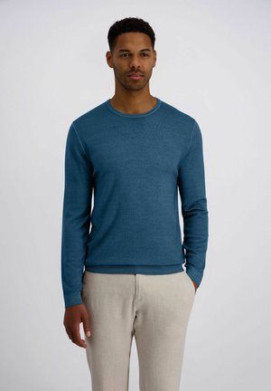 Mann steht mit den Händen in den Taschen, trägt einen blauen Pullover mit Rundhalsausschnitt und beige Hose vor einem einfarbigen weißen Hintergrund.
