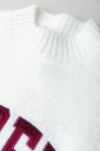 Hvit strikket genser med ribbestrikket krage og burgunder broderi. Teksturen ser myk ut med en avslappet passform og minimalt med detaljer.
