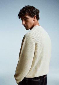 Cremig gestrickter Pullover mit Rundhalsausschnitt, langen Ärmeln und lockerer Passform, kombiniert mit dunklen Jeans mit farbiger Naht.