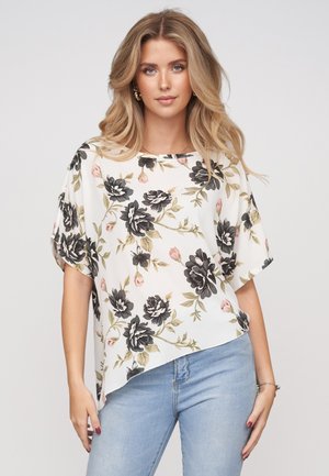 Kvinde med langt blond hår iført en hvid blomstret bluse med sorte og lyserøde blomster og lyseblå jeans mod en ensfarvet baggrund.