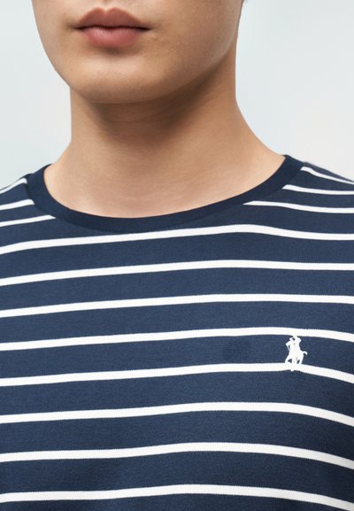 Νέος/νέα που φοράει μπλε ναυτικό και λευκό πουλόβερ με οριζόντιες ρίγες και μικρό λευκό κεντημένο λογότυπο με άλογο και αναβάτη στο στήθος.