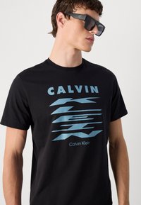 Juodas medvilninis marškinėliai su mėlynu grafiniu tekstu "CALVIN KLEIN" ir stilizuotomis linijomis. Dėvimi su tamsiais akiniais nuo saulės, pabrėžiantys įmantrų dizainą.