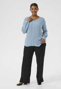 Blouse longue à manches longues bleu clair avec un col en V et un détail de boutons, associée à un pantalon noir à jambes larges et des chaussures noires. Tissu lisse, coupe décontractée.