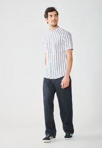 Camicia bianca a maniche corte con bottoni e righe verticali nere, abbinata a jeans in denim blu scuro e scarpe da ginnastica bianco e nere.
