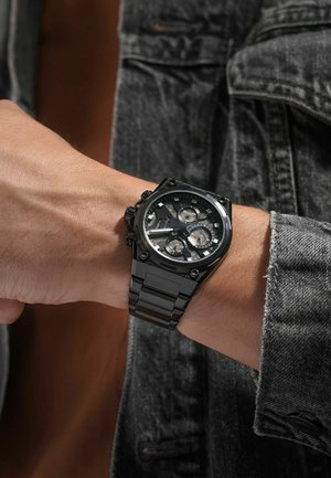 Montre en acier inoxydable noir avec un affichage multi-dial, dotée d'un bracelet texturé et de accents matériels prononcés. Portée sur un poignet contre du denim.