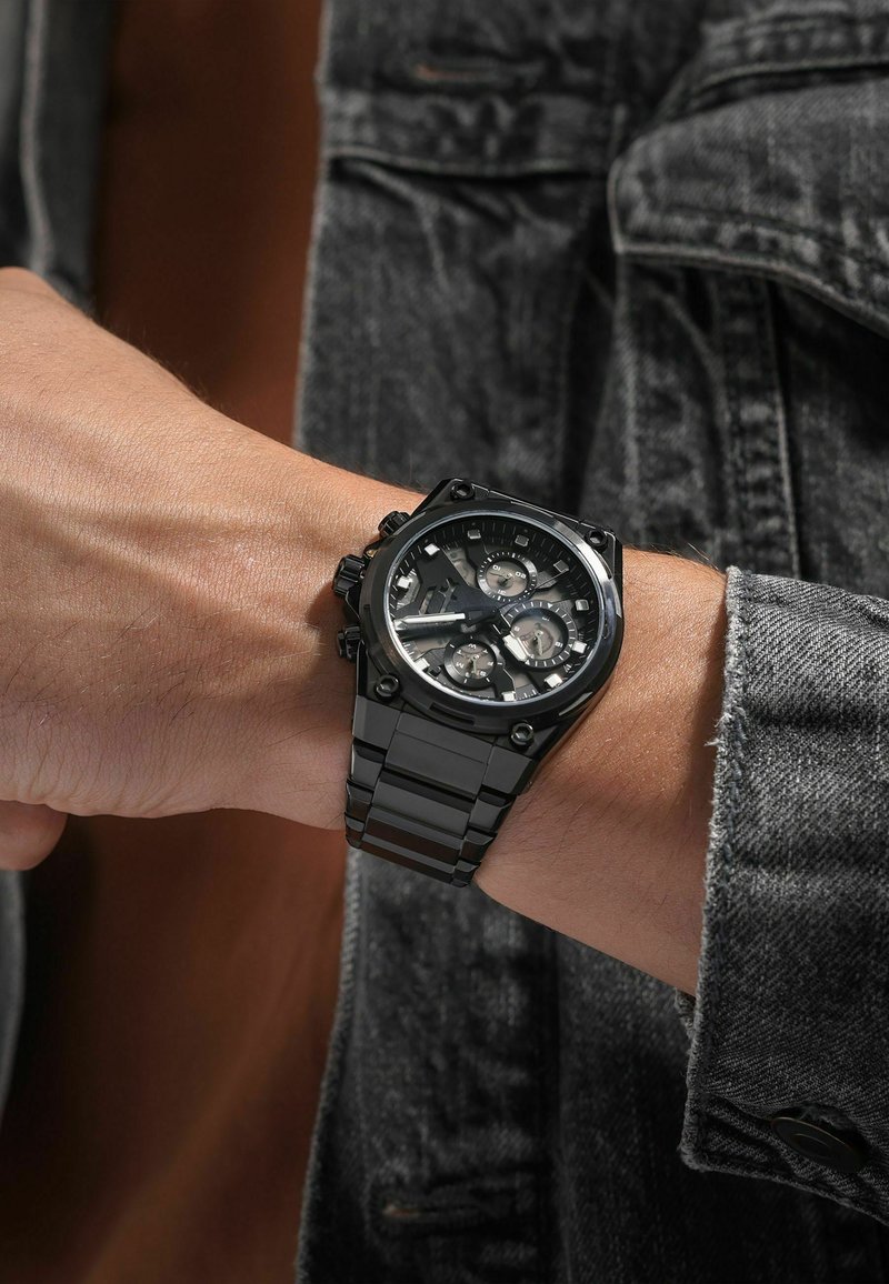 Orologio in acciaio inossidabile nero con quadrante multiplo, dotato di un cinturino testurizzato e di accentuazioni hardware prominenti. Indossato su un polso contro un paio di jeans.