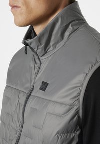 Graue gepolsterte Weste mit hohem Kragen, gestepptem Design, Frontreißverschluss und schwarzem Logo-Detail auf der Brust. Verfügt über minimale Designelemente.