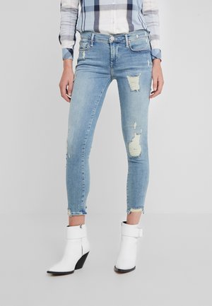 Femme portant un jean skinny bleu clair déchiré, des bottines blanches avec une boucle et une chemise à carreaux claire.