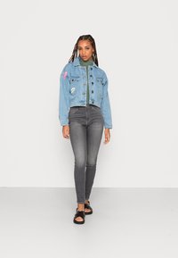Casaco de ganga em azul claro com botões e apliques, combinado com jeans skinny cinzentos e sandálias pretas. Modelo está posicionado contra um fundo branco.