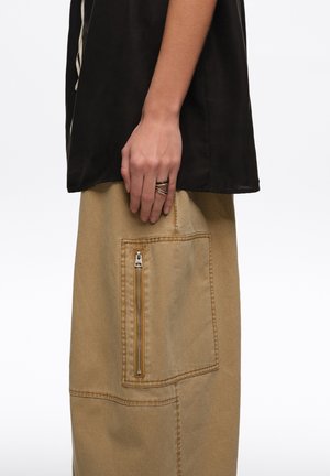 Persona con pantalones beige con un gran bolsillo lateral con cremallera y una camiseta negra, mostrando un anillo de varios aros en su mano izquierda.