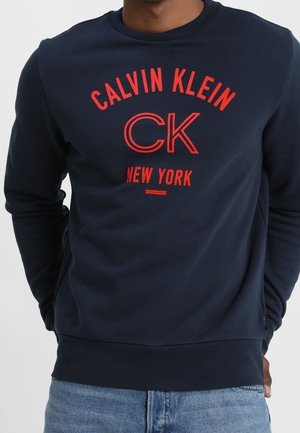 Sweatshirt marine à encolure ronde avec le logo rouge "Calvin Klein CK New York". Mélange de coton doux, coupe standard et manches longues.