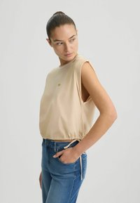Top beige senza maniche con scollatura rotonda, orlo arricciato e piccolo logo. Indossato con jeans in denim blu con tasca laterale.