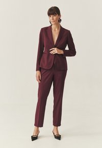 Bordeaux op maat gemaakt pak met een diepe V-hals blazer en slim-fit broek. Heeft strakke lijnen, een gladde textuur en puntige zwarte hakken.