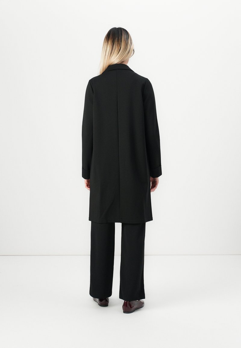 Frau mit langen blonden Haaren, die einer weißen Wand gegenübersteht, trägt einen langen schwarzen Mantel, schwarze Hose und dunkelrote Schuhe.