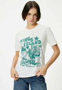 Hvid T-shirt med en turkis grafisk print og tekst om Kings Island forlystelsespark, der viser en stjerne, palmetræer og forlystelser.