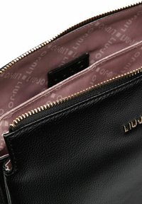 Zwarte leren tas met een getextureerd oppervlak, gouden rits en lichtroze voering met een herhalend logopatroon. Het interieur heeft twee compartimenten.