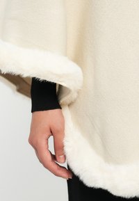Una mano poggiata vicino al bordo di un indumento in maglia crema con finitura in pelliccia sintetica bianca, indossato sopra una maglietta nera a maniche lunghe e pantaloni.