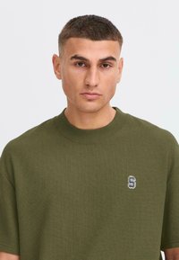 Olive green short-sleeve t-shirt with a textured knit pattern. It features a small embroidered logo on the left chest. Round neckline.
