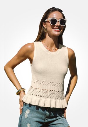Mujer con top blanco de punto sin mangas con bajo con volantes, gafas de sol, pendientes, pulsera y vaqueros azules desgarrados sonriendo con las manos en las caderas.