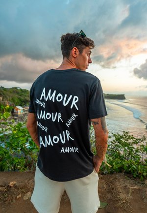 Črn bombažni T-majica z večkrat ponovljenim belim napisom "AMOUR" v različnih usmeritvah. Kratki rokavi, sproščen kroj, okroglo ovratnik.