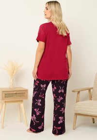 T-shirt rouge à manches courtes associé à un pantalon noir avec des motifs de nœuds roses. Le modèle se tient devant un fond neutre à côté d'une table en bois.