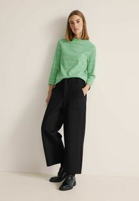 Maglione verde a coste con maniche lunghe, abbinato a pantaloni neri larghi con tasche e cintura con bottoni, indossato con scarpe nere.