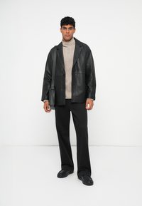 Camicia overshirt in pelle nera con una finitura testurizzata, abbinata a un dolcevita beige a maglia cable-knit, pantaloni larghi neri e scarpe slip-on nere.
