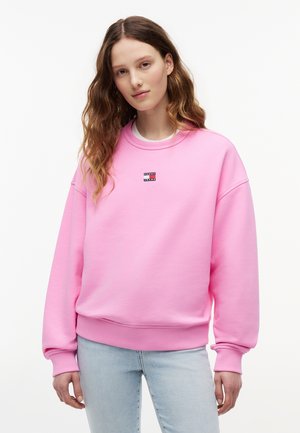 Νεαρή γυναίκα με κυματιστά μαλλιά που φοράει έντονη ροζ φούτερ και ανοιχτόχρωμο μπλε τζιν μπροστά σε απλό λευκό φόντο.