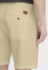 Shorts en coton beige avec un ourlet roulé, des poches latérales et une poche arrière avec un patch en cuir marron portant la marque et un détail de bouton.