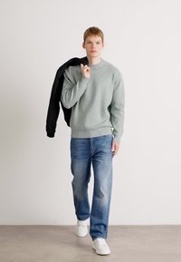 Junger Mann, der mit einem hellgrauen Pullover, blauen Jeans und weißen Sneakers nach vorne geht, während er eine schwarze Jacke über der Schulter hält.
