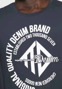 Navy blue cotton t-shirt featuring white circular graphic with “Tom Tailor Denim” logo, design elements, and established date text.