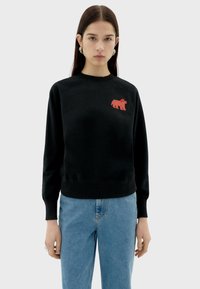 Zwarte sweater met geribbelde manchetten en onderkant, met een rood gestreept berenlogo op de borst. Gecombineerd met lichtblauwe jeans.