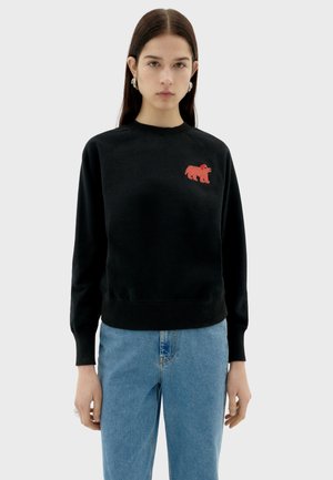 Schwarzer Pullover mit gerippten Bündchen und Saum, mit einem roten gestreiften Bärenlogo auf der Brust. Kombiniert mit hellblauen Jeans.