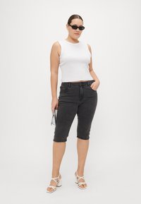 Top corto blanco, pantalones cortos de mezclilla negros hasta la rodilla, tacones blancos con tiras y un pequeño bolso plateado. La modelo lleva gafas de sol y tiene el cabello liso.
