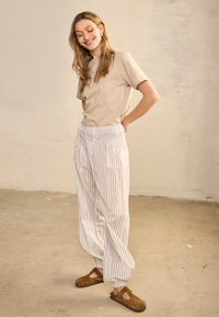 Mosolygó nő hosszú hajjal, bézs pólóban, laza fehér nadrágban függőleges csíkokkal és barna papucsban, beltéren állva.