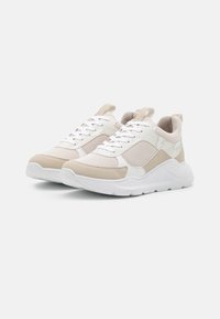 Zapatos deportivos en color beige y blanco, con material de malla y ante, lengüeta acolchada y detalles texturizados en los laterales, que cuentan con una suela blanca gruesa.