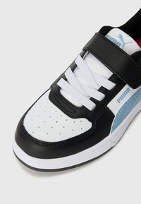 Chaussure de sport noir et blanc avec une tige en cuir, fermeture à lacets et Velcro, bout perforé, accents bleus et semelle en caoutchouc.