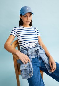 Chemise à rayures blanches et bleues à manches courtes, casquette bleu clair, et veste en jeans nouée autour de la taille, assis sur une chaise en bois devant un fond pastel.