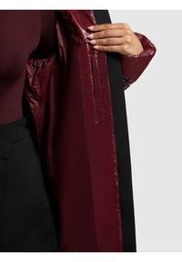 Osoba oblečená v černých kalhotách a tmavě burgundském topu, držící otevřený dlouhý černý kabát, který odhaluje lesklou burgundskou podšívku a vnitřní kapsu.