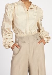 Blouse crème à rayures verticales, avec des manches smockées et un col ; associée à un pantalon beige taille haute avec des poches latérales et des détails de boutons.
