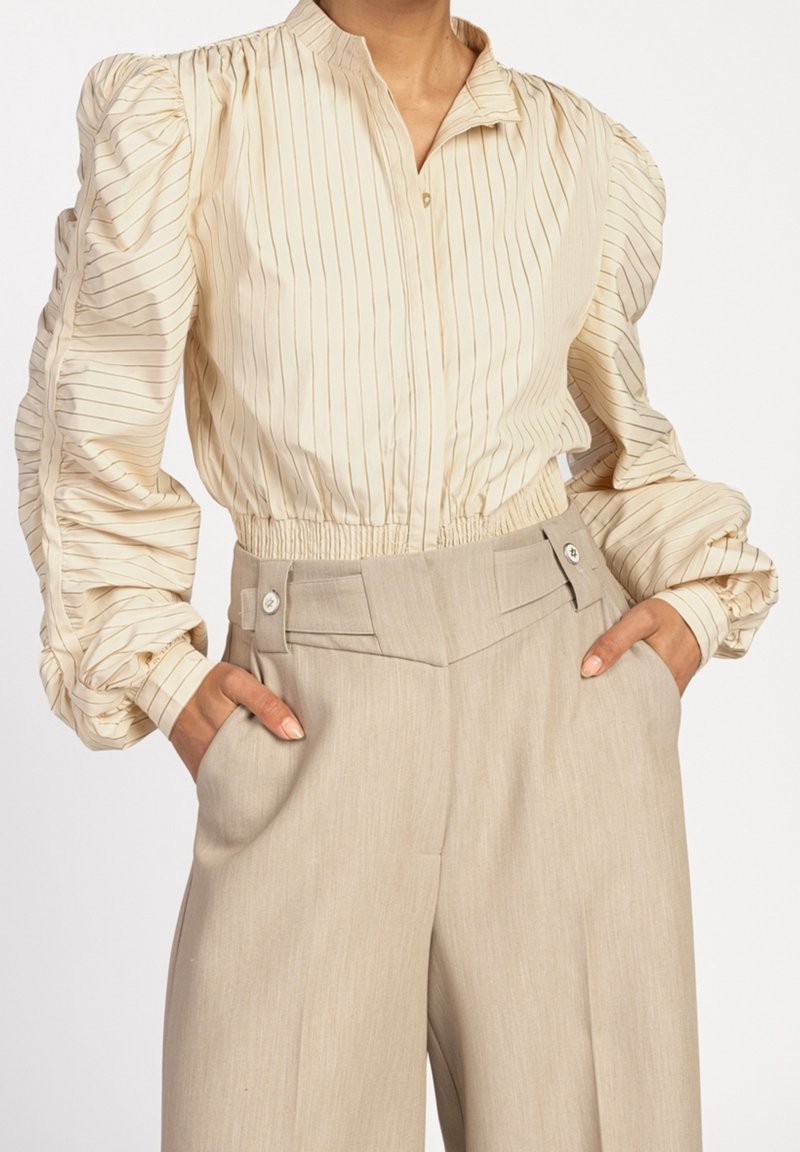 Blouse crème à rayures verticales, avec des manches smockées et un col ; associée à un pantalon beige taille haute avec des poches latérales et des détails de boutons.