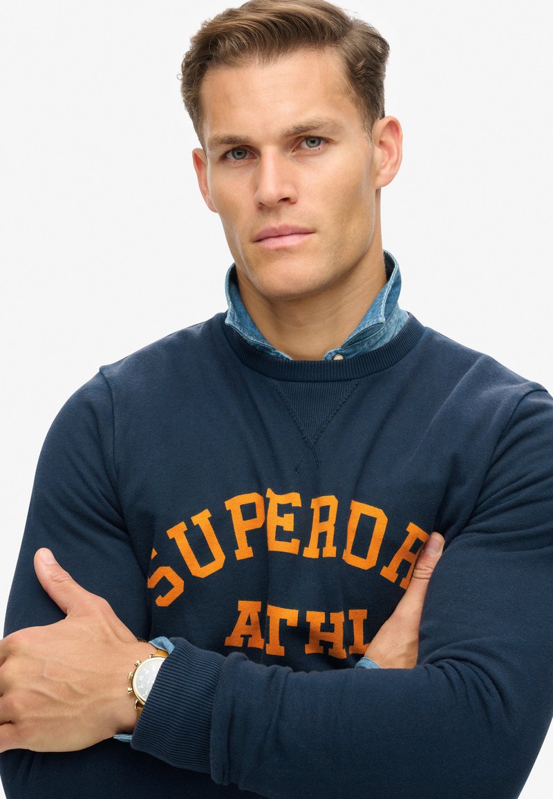 Marineblaues Sweatshirt mit "SUPERDRY ATHL" in Orange. Strukturierter Stoff, gerippte Bündchen, über einem hellen Jeanshemd getragen, Uhr am Handgelenk.