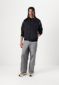 Chaqueta ligera negra con botones a presión, pantalones cargo grises con bolsillos, combinados con zapatillas beige sobre un fondo neutro.