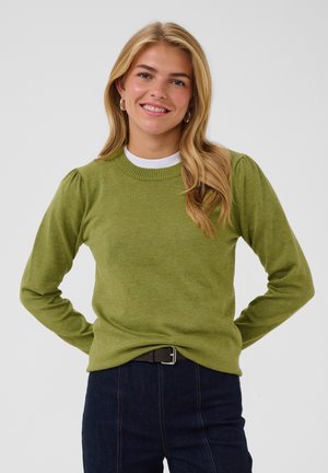 Jeune femme aux cheveux blonds portant un pull vert et un jean foncé, souriante avec les mains derrière le dos sur un fond uni.
