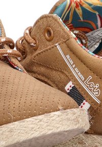 Zapatos de lona marrones con perforaciones y suela de yute trenzada. Presentan un forro floral colorido y una etiqueta de logo en el lado exterior.