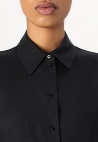 Camicia nera con colletto realizzata in tessuto texturizzato, con chiusura a bottoni neri e colletto liscio.